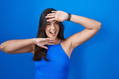 Hispanic woman standing over blue background smiling cheerful playing peek a boo with hands showing face. surprised and exited 