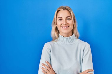Young caucasian woman standing over blue background happy face smiling with crossed arms looking at the camera. positive person. 