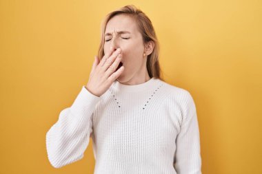 Sarı arka planda beyaz süveter giyen genç beyaz kadın ağzıyla esnemekten sıkılmış. Huzursuz ve uykulu.. 