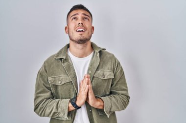 Tecrit edilmiş arka planda duran genç İspanyol adam elleri bağlı yalvarıyor ve dua ediyor. Yüzünde umut ifadesi var. Çok duygusal ve endişeli. Yalvarıyor. 