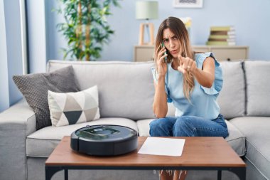 Evde vakumlu robotla oturan genç İspanyol kadın telefonla konuşuyor. Kamerayı işaret ediyor ve kendine güveni tam bir jest yapıyor. 