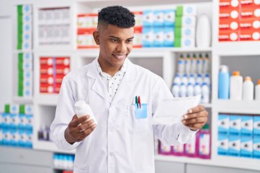 Young latin man pharmacist holding pills bottle reading prescription at pharmacy