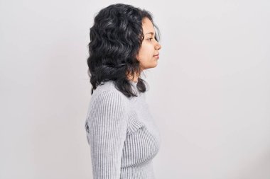 Esmer, koyu renk saçlı bir kadın. Arka planda duruyor. Yan tarafa bakıyor, rahat bir poz veriyor.. 