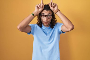 Young hispanic man standing over yellow background doing funny gesture with finger over head as bull horns 