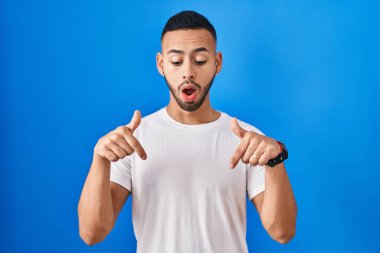 Mavi arka planda dikilen genç İspanyol adam parmaklarını göstererek reklamını yapıyor, şaşırmış yüz ve ağzını açıyor. 