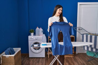 Çamaşır odasında ütü yapan genç esmer kadın kameraya seksi bir ifadeyle bakıyor, neşeli ve mutlu bir yüzle.. 