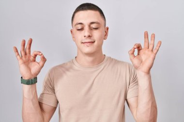 İzole edilmiş arka planda duran genç adam rahatlamış ve parmaklarıyla meditasyon hareketi yaparken gözleri kapalı gülümsüyor. yoga konsepti. 