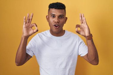 Young hispanic man standing over yellow background looking surprised and shocked doing ok approval symbol with fingers. crazy expression 