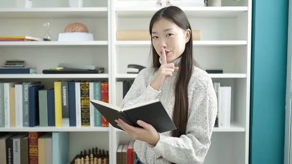 Young chinese woman student reading book doing silence gesture at library university