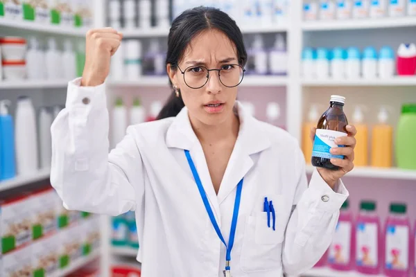 Young hispanic woman working at pharmacy drugstore holding syrup annoyed and frustrated shouting with anger, yelling crazy with anger and hand raised