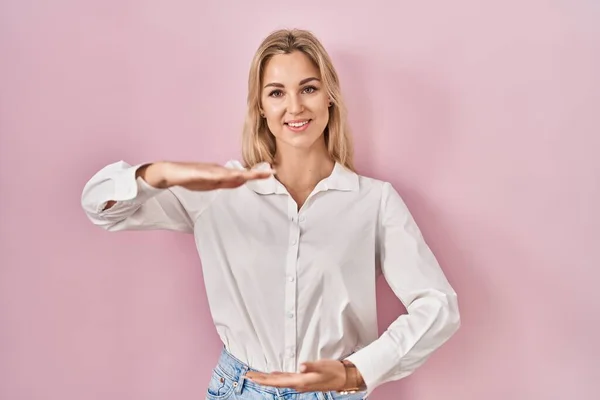 年轻的高加索女人 穿着宽松的白色衬衫 背景是粉色的 手部有大而大的标志 尺寸是符号 微笑着看着摄像机 计量概念 — 图库照片