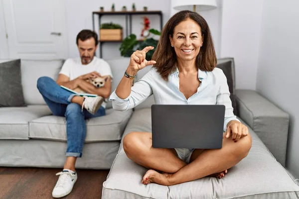 Hispanic middle age couple at home, woman using laptop smiling and confident gesturing with hand doing small size sign with fingers looking and the camera. measure concept.
