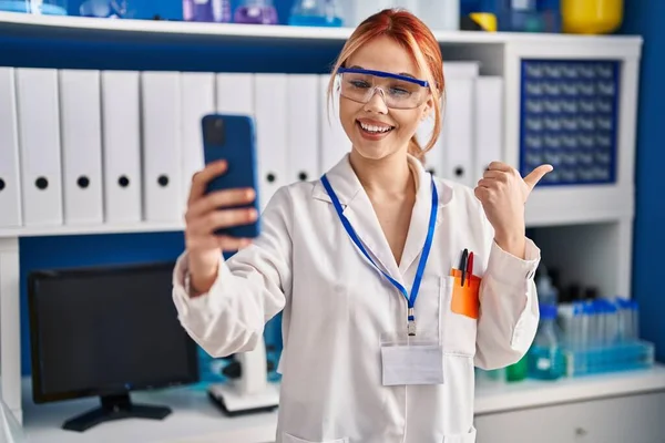 在科学家实验室工作的年轻的高加索女人用智能手机打了一个视频电话 手指指向旁边 笑着张开了嘴 — 图库照片