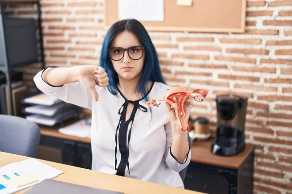 Young girl with blue hair holding model of female genital organ at the office complaining for menstruation pain with angry face, negative sign showing dislike with thumbs down, rejection concept