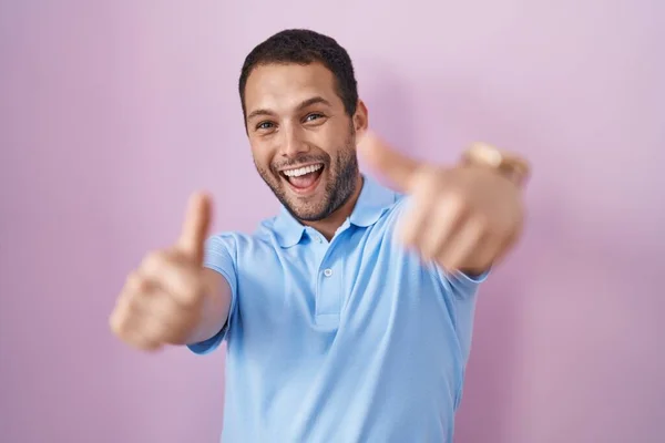 ピンクの背景に立っているヒスパニック系の男性は 手で積極的なジェスチャーを行うことを承認し 親指を笑顔と成功のために幸せ 勝者のジェスチャー — ストック写真
