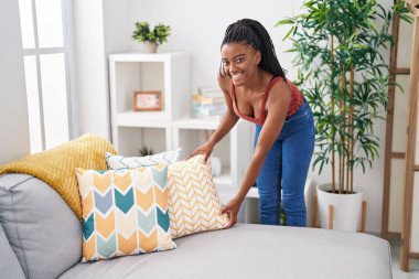 Afro-Amerikalı kadın gülümsüyor. Evdeki kanepeyi organize ediyor.