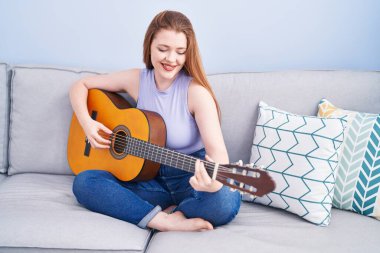 Young redhead woman playing classical guitar sitting on sofa at home