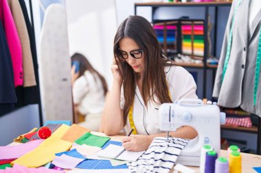 Young beautiful hispanic woman tailor talking on smartphone writing on notebook at clothing factory