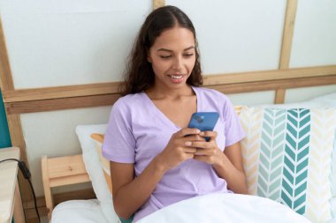 Young african american woman using smartphone sitting on bed at bedroom