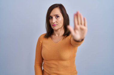 Orta yaşlı esmer kadın turuncu kazak giyip avuç içiyle şarkı söylemeyi kesiyor. Yüzünde olumsuz ve ciddi bir jest olan uyarı ifadesi. 