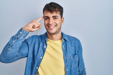 Young hispanic man standing over blue background smiling pointing to head with one finger, great idea or thought, good memory 