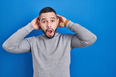 Mavi arka planda dikilen İspanyol adam deli ve elleri başının üstünde korkmuş ve ağzı açık şoktan korkmuş. 