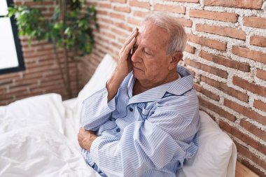 Orta yaşlı, kır saçlı, stresli bir adam yatak odasında oturuyor.