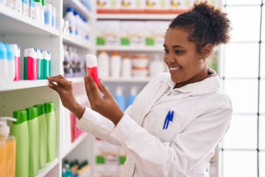 African american woman pharmacist smiling confident holding toothpaste of shelving at pharmacy