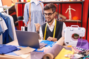 Dövmeleri olan İspanyol bir genç terzi terzi, terzi odasında dizüstü bilgisayar kullanıyor başparmağını yana uzatıyor ve ağzı açık gülümsüyor. 