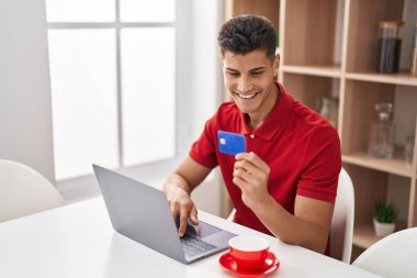 Young hispanic man using laptop and credit card sitting on table at home