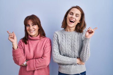 Anne ve kızı mavi arka planda, yüzünde kocaman bir gülümsemeyle, kameraya el ile işaret ederek duruyorlar.. 