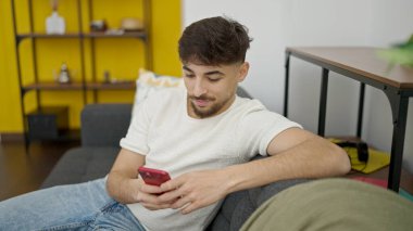 Young arab man using smartphone sitting on sofa at home