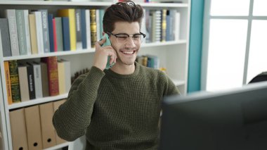 Young hispanic man student talking on smartphone using computer at library university