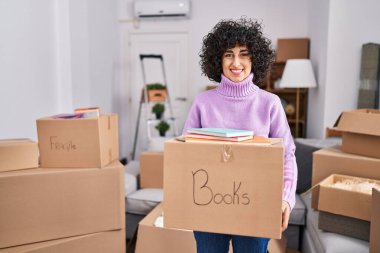 Kıvırcık saçlı genç esmer kadın elinde karton bir kutuyla yeni evine taşınıyor yüzünde mutlu ve havalı bir gülümsemeyle gülümsüyor. diş gösterme. 