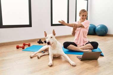 Genç beyaz kadın spor merkezinde köpekle internetten esneme dersi alıyor.