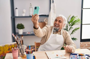 Orta yaşlı, gri saçlı, gülümseyen bir sanatçı sanat stüdyosundaki akıllı telefondan özçekim yapıyor.