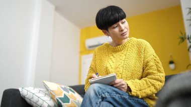 Young chinese woman writing on notebook sitting on sofa at home
