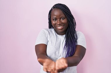 Pembe arka planda duran Afrikalı genç bir kadın avuçlarını birbirine uzatarak gülümsüyor ya da el hareketi yapıyor. Bekleme ve koruma 