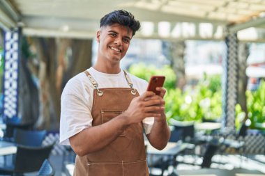Genç İspanyol garson, kahvecinin terasında akıllı telefon kullanarak gülümsüyor.