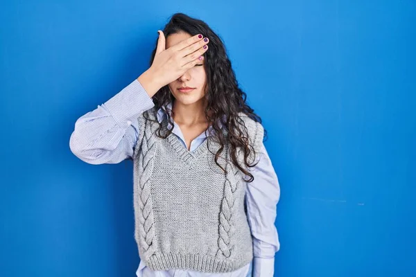 Genç esmer kadın mavi arka planda dikiliyor. Elle gözlerini kapatıyor, ciddi ve üzgün görünüyor. Görme, saklama ve reddetme konsepti 