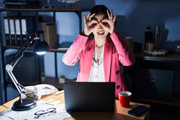 Chinese young woman working at the office at night doing ok gesture like binoculars sticking tongue out, eyes looking through fingers. crazy expression.