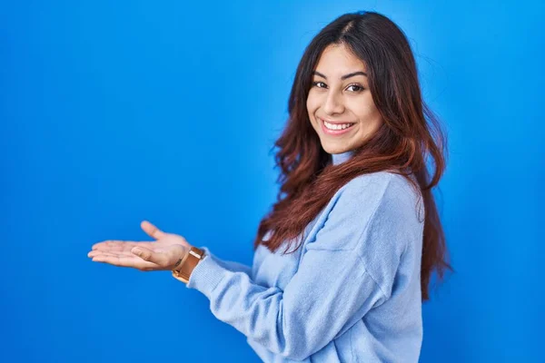 西班牙裔年轻女子站在蓝色的背景旁边 双手张开手掌 展示复制的空间 呈现广告的笑容让人兴奋而兴奋 — 图库照片