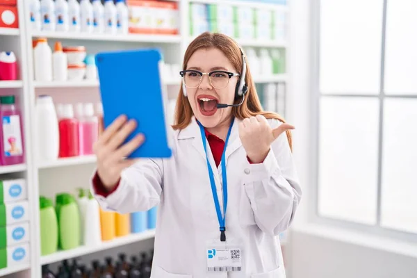 在药店工作的红头发女人用平板电脑打了一个视频电话 手指指向旁边 笑着张开了嘴 — 图库照片