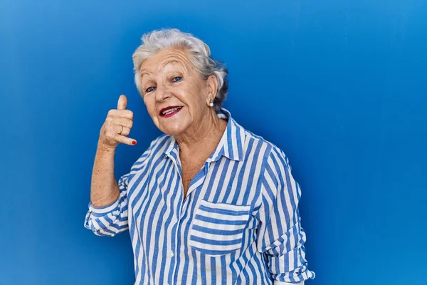 白髪のシニア女性が青い背景の上に立ち 電話で話すような手や指で電話のジェスチャーをして笑っています コミュニケーションの概念 — ストック写真
