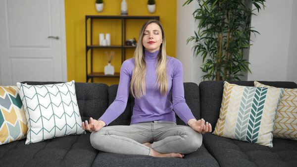 Young blonde woman doing yoga exercise sitting on sofa at home