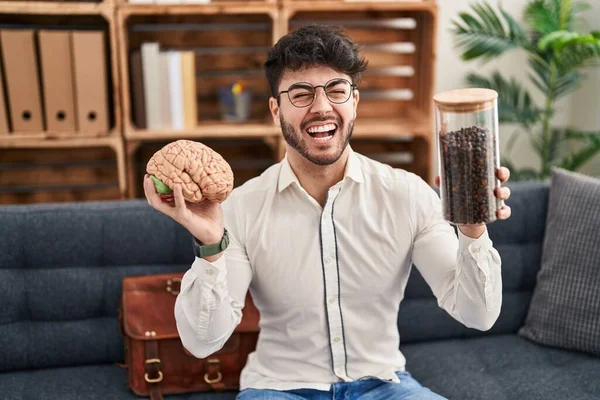 Hispanic man with beard working at therapy office holding brain and coffee beans angry and mad screaming frustrated and furious, shouting with anger. rage and aggressive concept.