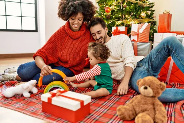 Couple and daughter playing with toys sitting by christmas tree at home