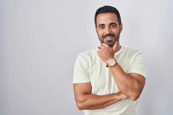 Hispanic man with beard standing over isolated background looking confident at the camera smiling with crossed arms and hand raised on chin. thinking positive.