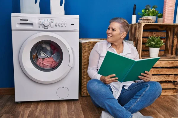 洗濯室で洗濯機を待っている本を読んでいる中年女性 — ストック写真
