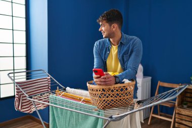 Genç İspanyol adam çamaşır odasında çamaşır ipine elbise asarak akıllı telefon kullanıyor.